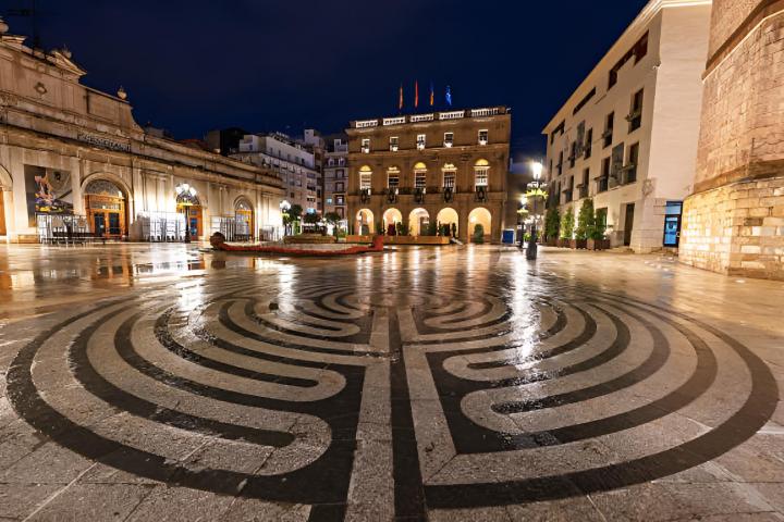 La Plaza Mayor de Castellón de la Plana, con un mosaico circular laberíntico y el Ayuntamiento y el Teatro Principal iluminados por la noche.
