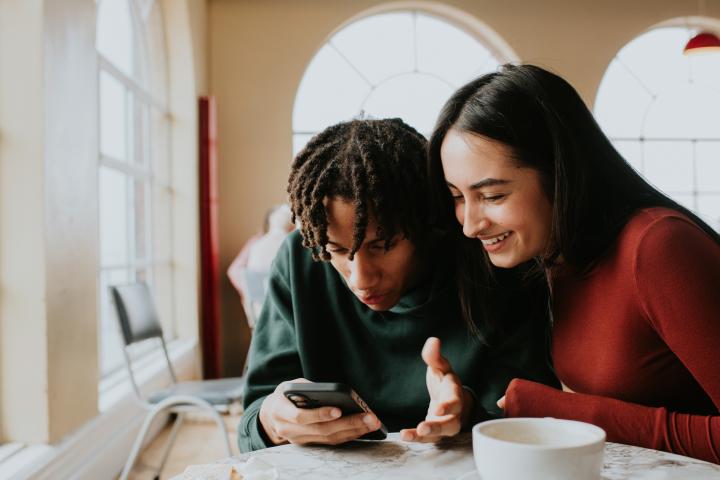 alt="alt="Dos jóvenes adultos sentados cerca en una mesa de café, compartiendo un smartphone y reaccionando con interés.""
