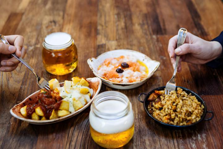 Una mesa de bar llena de tapas españolas y dos cervezas.