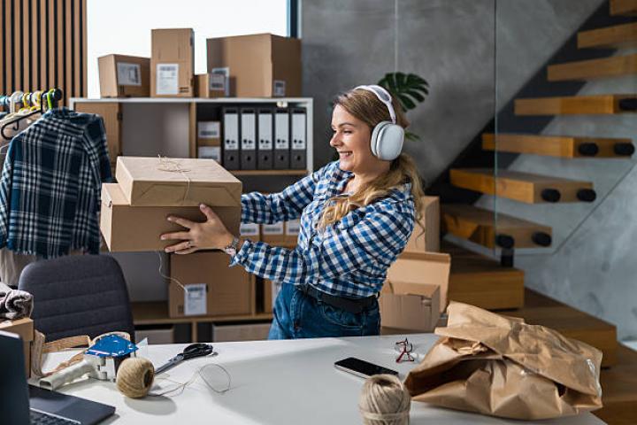A cheerful woman wearing headphones packs cardboard boxes in a modern office filled with supplies and office gear. Perfect for topics on shipping, e-commerce, packaging, and small business.