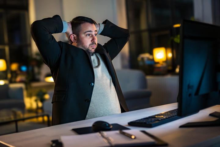 Un hombre de mediana edad, vestido de manera profesional, se sienta estresado en una oficina poco iluminada por la noche, usando una computadora de escritorio con papeles sobre el escritorio.