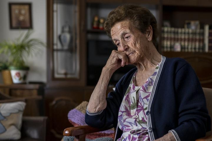 Mujer mayor sentada sola en un sofá, con la mirada triste, pensando en problemas, recordando momentos de su vida, extrañando a sus hijos y sintiéndose sola.