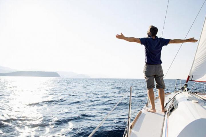 Un hombre de pie en la proa de un yate con los brazos extendidos y mirando al horizonte