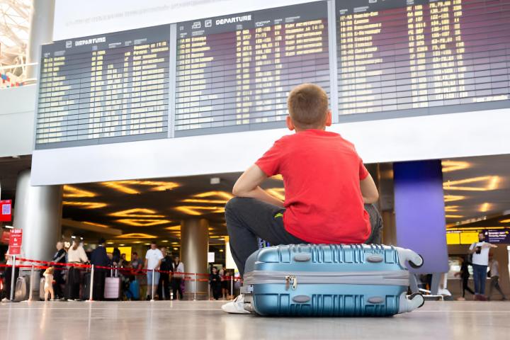 Un niño solitario sentado en una maleta en el aeropuerto mirando el tablero de llegadas