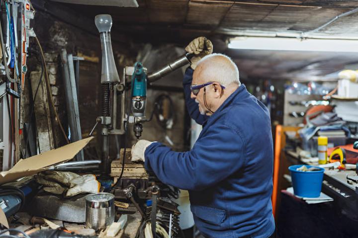 Un hombre anciano usando el taladro en su taller con gafas y guantes de protección