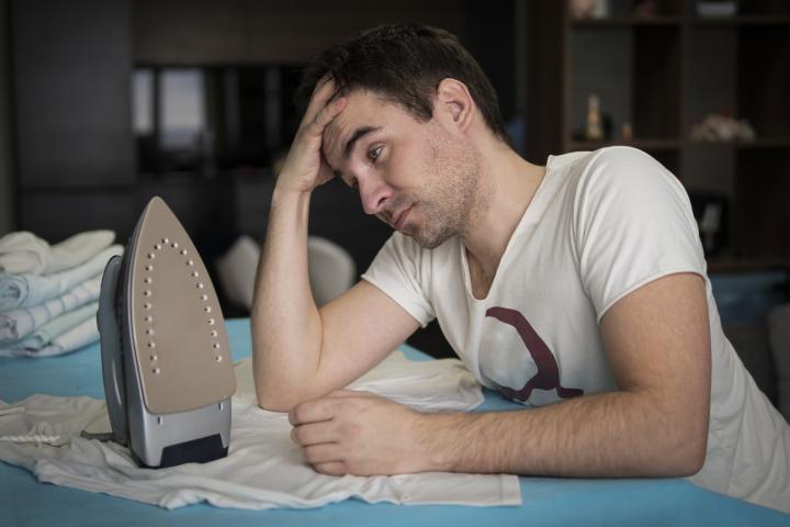 Un joven plancha su ropa en casa, con el interior de la vivienda de fondo.