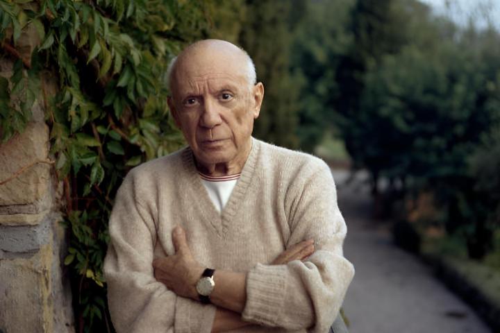 Pablo Picasso de pie junto a un helecho verde, con los brazos cruzados y vistiendo un suéter de cachemir.