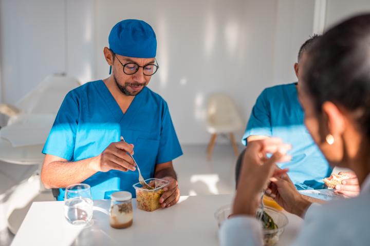 Un médico con uniforme azul almuerza mientras conversa con sus compañeras en una sala de descanso de un hospital bien iluminada, lo que representa un momento de descanso en un entorno médico.