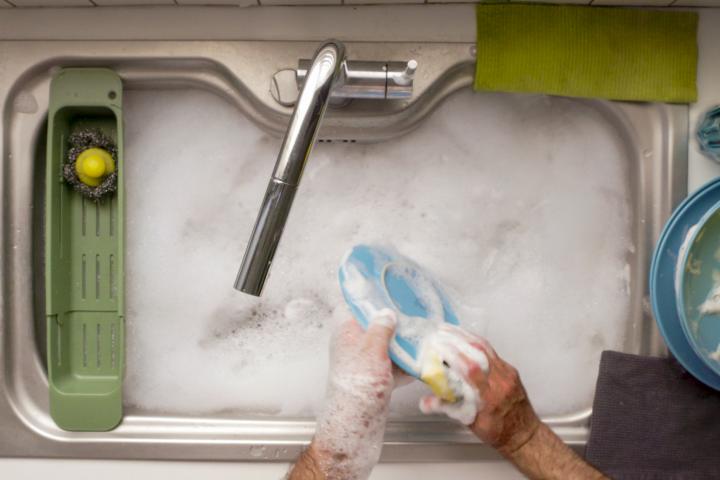 Vista superior de unas manos fregando platos en un fregadero lleno de jabón y espuma. Una colorida variedad de utensilios y vajilla, que incluye un cuenco azul y tazas amarillas, realza el ambiente animado.