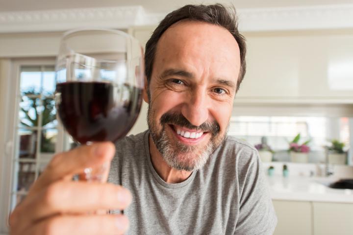 Hombre de mediana edad bebiendo una copa de vino con rostro feliz, de pie y sonriendo con confianza mostrando los dientes.