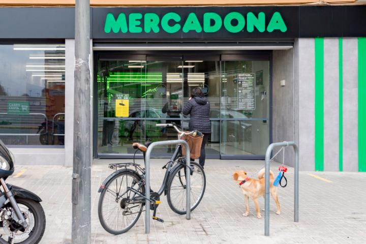 : Una mujer entra en el supermercado Mercadona, mientras un perro está atado frente a la puerta.
