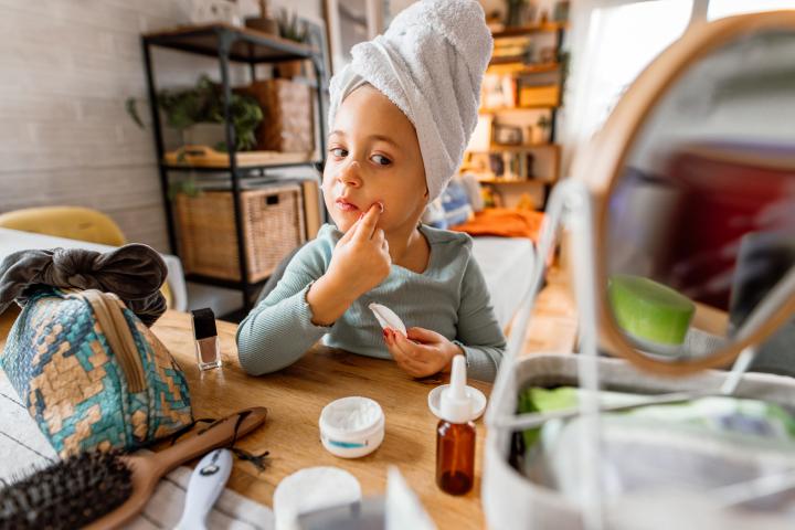 Una niña pequeña sonriente, con una toalla enrollada alrededor de la cabeza, se mira al espejo mientras se aplica crema hidratante en la cara.