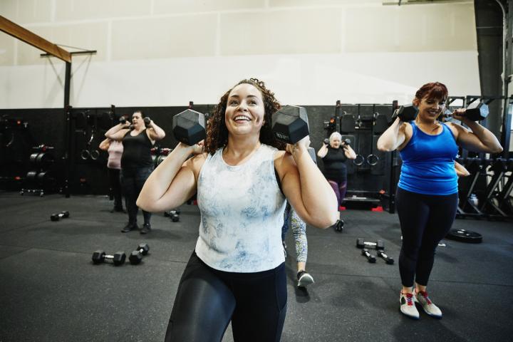 Mujer sonriente haciendo zancadas con mancuernas durante una clase de fitness en el gimnasio.