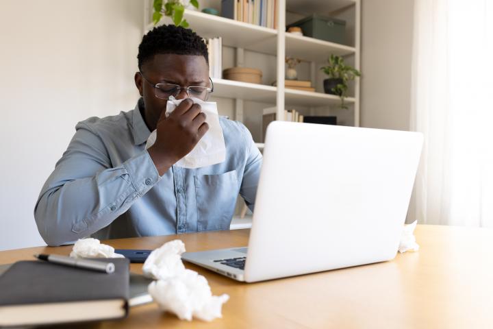 Un hombre adulto se suena la nariz con un pañuelo mientras trabaja a distancia en su portátil.