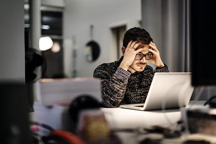 Un joven hombre de negocios, con gafas y aspecto informal, sentado frente a su portátil, con la cabeza entre las manos