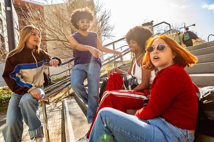 Four friends in casual street style sit on city steps, laughing and chatting in sunny, vibrant urban surroundings, showing diverse gen z friendship and carefree leisure