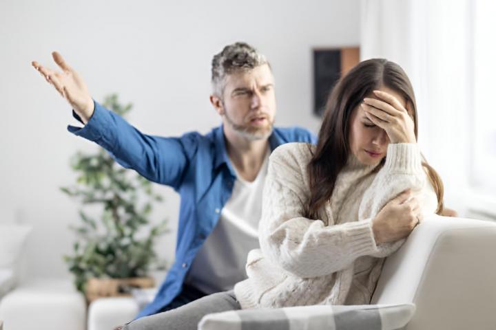 Un marido gritando a su pareja en el sofá de casa, quien lo ignora y se lleva las manos a la cabeza con frustración.