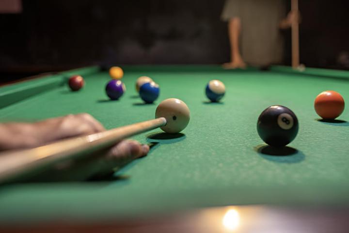 Primer plano de un taco de billar golpeando la bola blanca sobre una mesa verde, con bolas de colores al fondo, que simbolizan la concentración, la habilidad y la precisión en el juego.