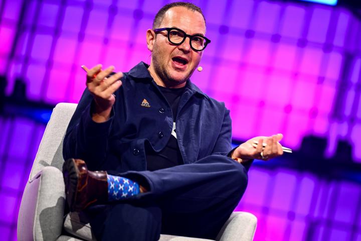 Cory Doctorow dando una charla en Lisboa, Portugal, en 2025. El plano está inclinado, tiene las piernas cruzadas, los brazos abiertos, gesticulando, y el fondo es un panel LED rosa.