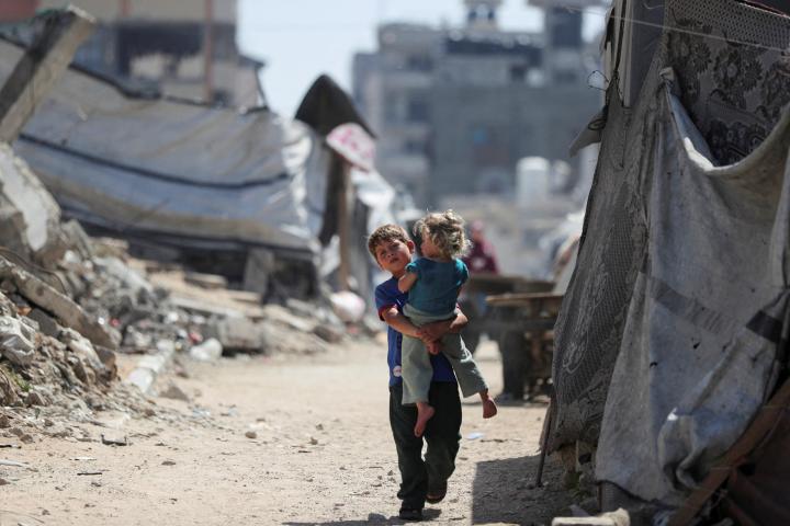 Un menor palestino, cargando a una niña, camina por una zona dañada entre los escombros de edificios residenciales en la ciudad de Gaza, el 11 de mayo de 2026.