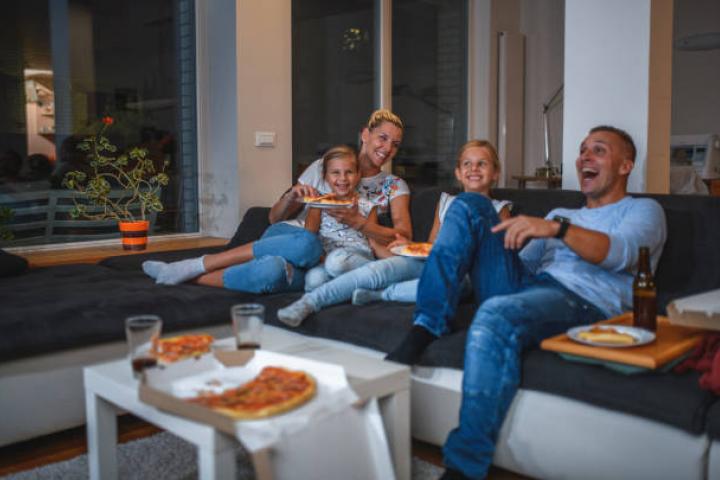Caucasian Family with two girls is sitting on a sofa in living room, watching TV and eating pizza.