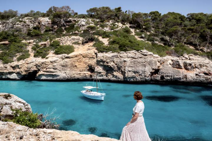 Una mujer contemplando el mar en una pequeña playa virgen rodeada de árboles con un yate en Mallorca.
