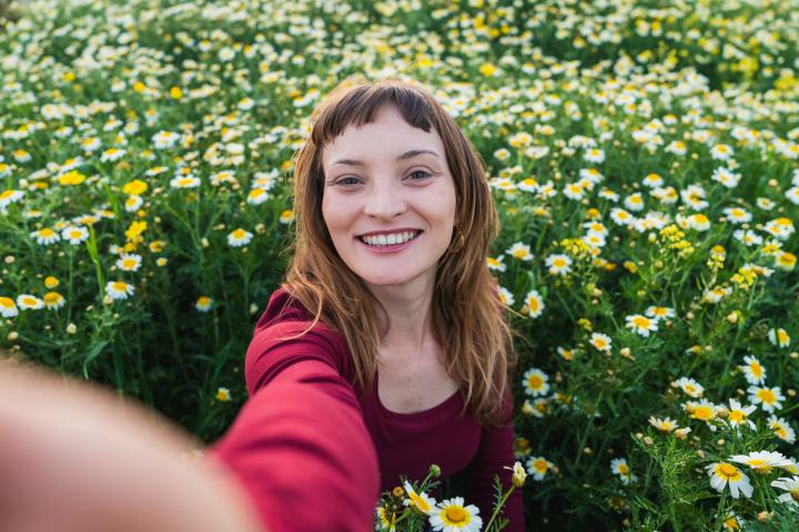 Una mujer sonriendo mientras se toma una fotografía tipo selfie en un campo lleno de margaritas