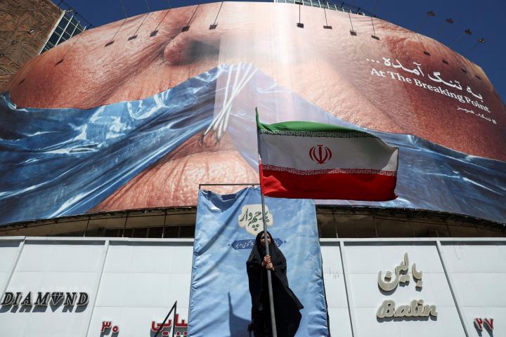 Una mujer sostiene una bandera iraní ant una valla antiestadounidense que muestra al presidente Donald Trump amordazado por el estrecho de Ormuz, en Teherán, el 25 de mayo de 2026.