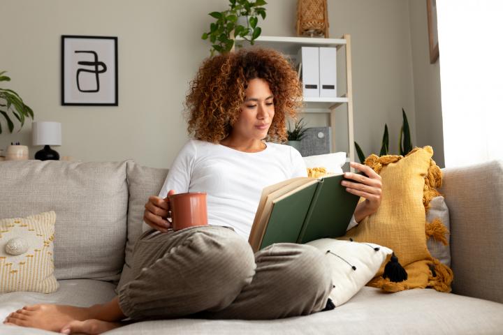 Mujer joven leyendo un libro en casa, tomando té sentada en el sofá de una acogedora sala de estar.
