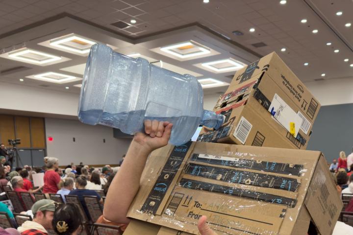 Un hombre disfrazado con varias cajas de Amazon bebe de un barril de agua, en una protesta contra la construcción de centros de datos en EEUU