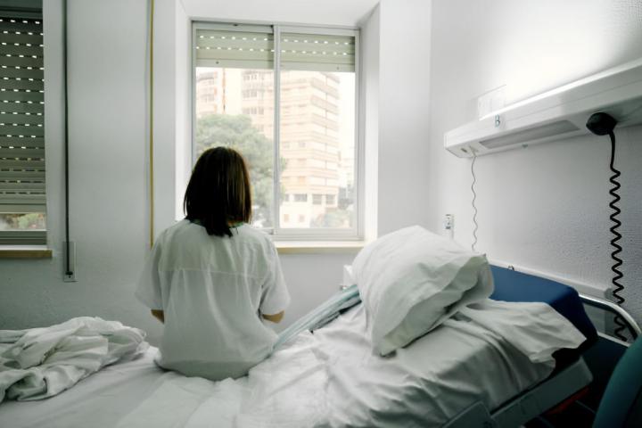 Una mujer enferma sola sentada en la cama de una habitación de hospital mirando por la ventana vista desde atrás