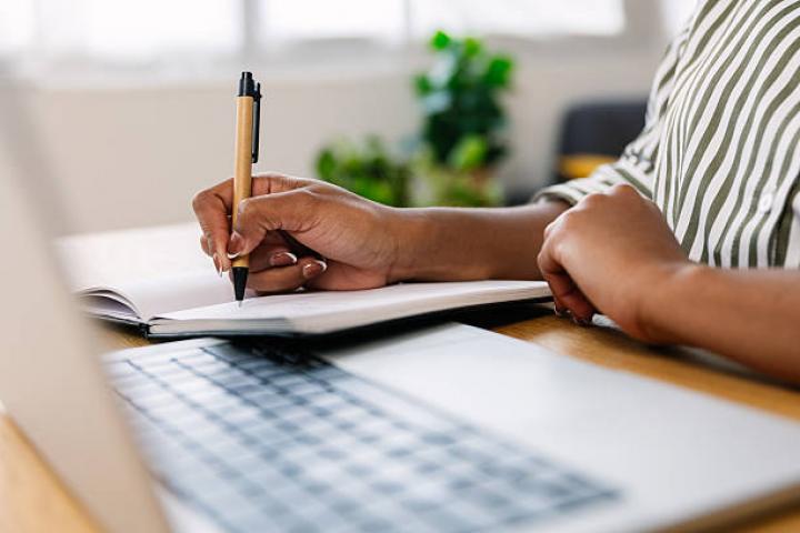 Unrecognizable businesswoman taking notes in a notebook while working on her laptop in a cozy home office, balancing productivity and comfort. Business and telework concept