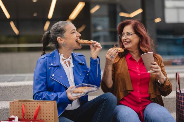 Two senior Caucasian women with shopping bags rest on the stairs while eating a cookie and drinking coffee after a day of shopping in a mall.
