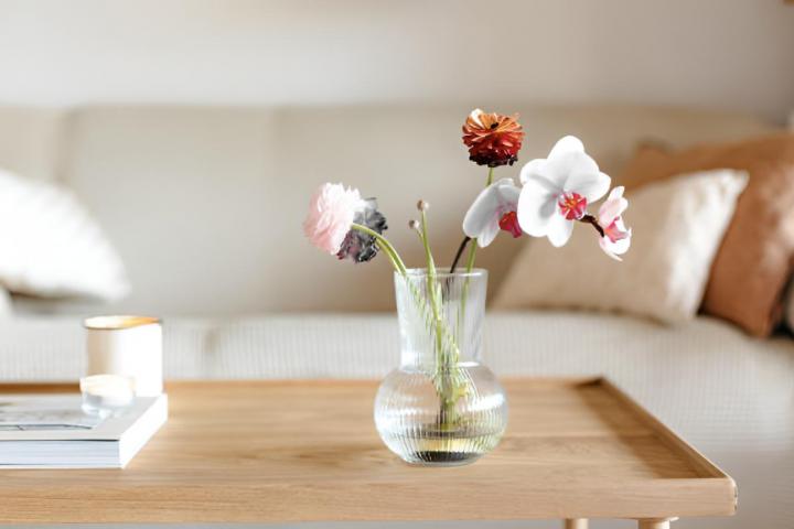 Un salón con una mesa de café y madera, flores en un jarrón de cristal y un sofá con cojines al fondo.