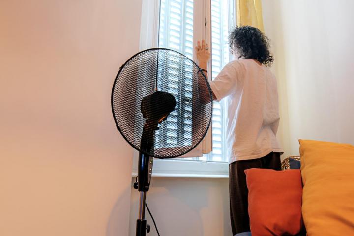 Una mujer cerrando las ventanas de su casa por calor mientras tiene encendido un ventilador