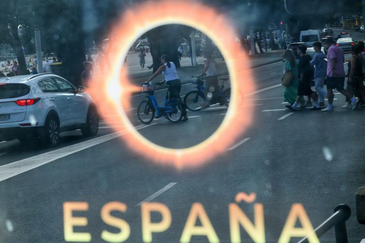 Gente pasea en A Coruña y se ve reflejada sobre un cartel que anuncia el gran eclipse solar total del 12 de agosto