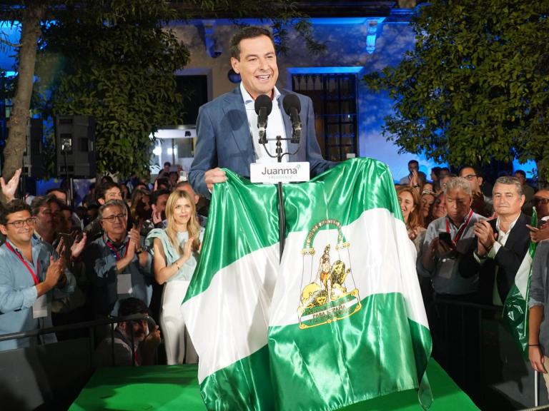 Subido en un escenario, rodeado de simpatizantes del PP, Juanma Moreno Bonilla sostiene una bandera de Andalucía