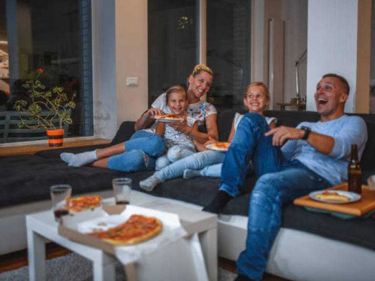 Caucasian Family with two girls is sitting on a sofa in living room, watching TV and eating pizza.
