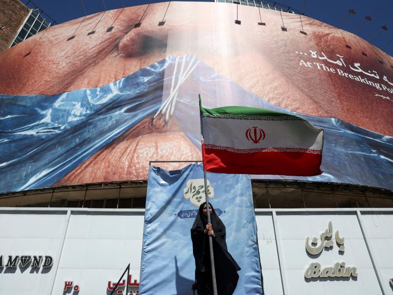 Una mujer sostiene una bandera iraní ant una valla antiestadounidense que muestra al presidente Donald Trump amordazado por el estrecho de Ormuz, en Teherán, el 25 de mayo de 2026.